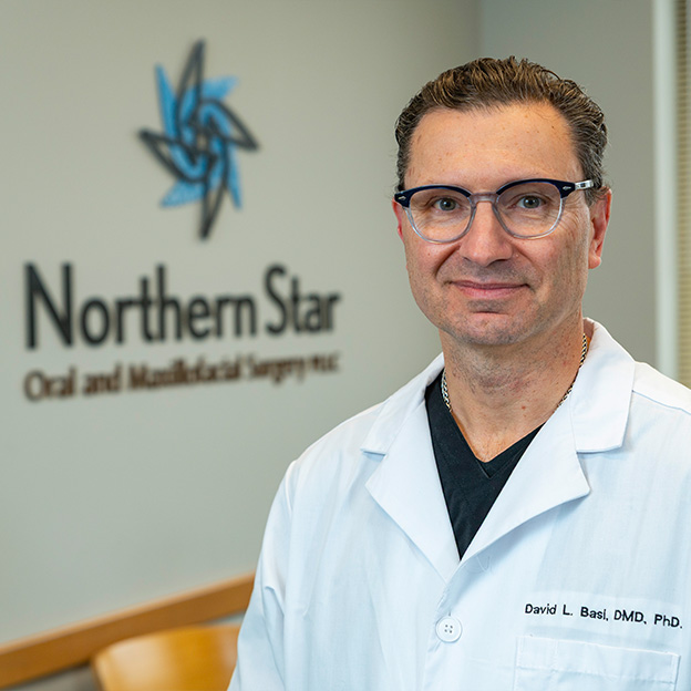 The image shows a man wearing glasses, standing in front of a sign with the text Northern Star, smiling at the camera. He appears to be a professional, possibly a dentist or medical practitioner, given his attire and the context provided by the sign behind him.