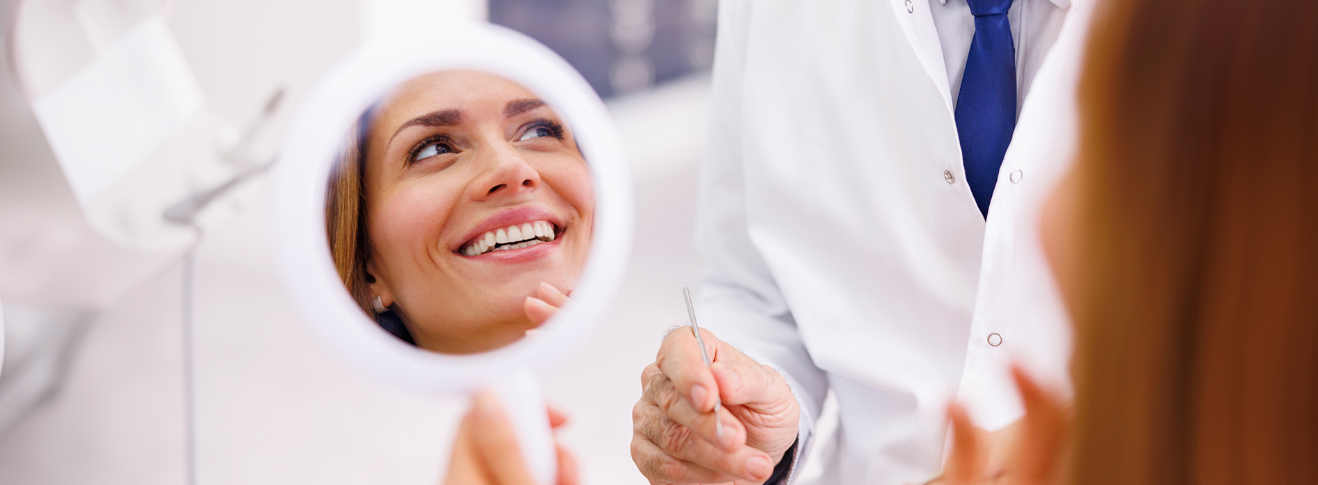 The image depicts a woman sitting in front of a mirror with a dentist, who is smiling at her reflection.