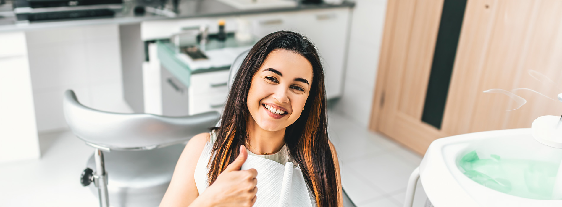 This is a photograph of a woman giving a thumbs-up gesture while seated at a salon chair, smiling towards the camera.
