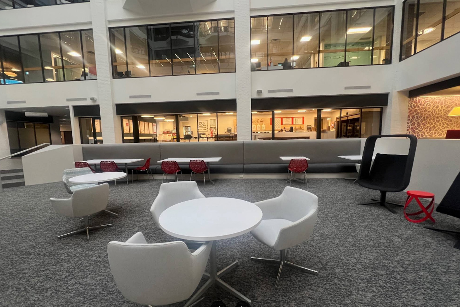 The image depicts a modern, spacious office interior with contemporary furniture, including chairs, tables, and seating areas, arranged around a central open space with natural light from large windows.