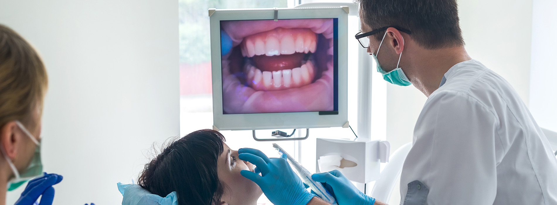 The image displays a dental setting where a dentist is examining a patient s mouth using a large screen for visualization, with two other individuals present, one of whom appears to be assisting or observing.