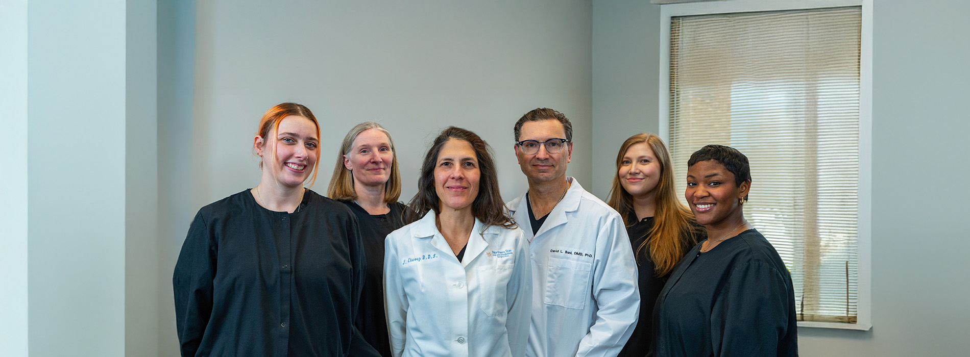 The image shows a group of individuals posing together, likely professionals given their attire, which includes white lab coats and stethoscopes. They are standing in front of a plain background with a window on the right side.