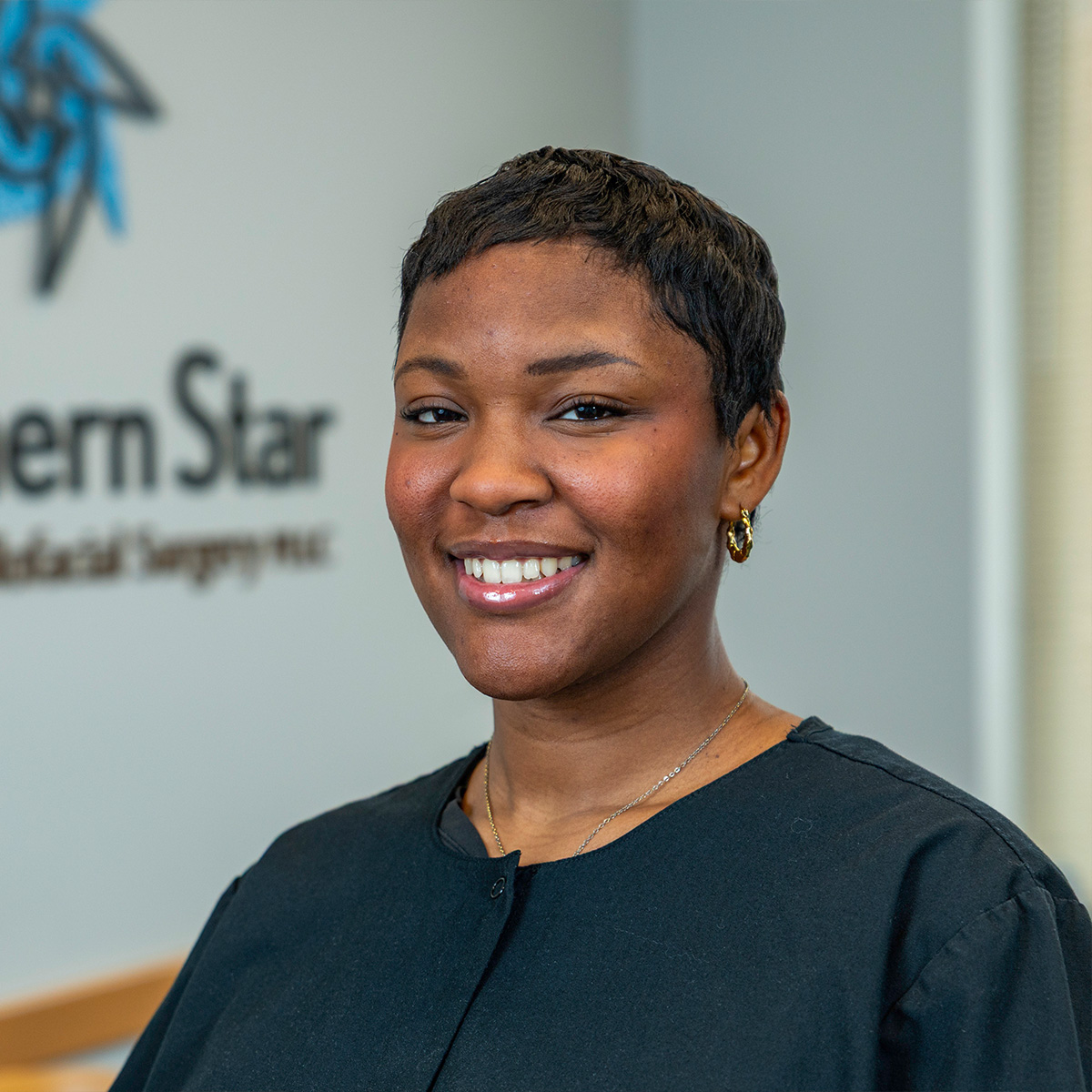 The image shows a woman standing indoors with a smiling expression, wearing a black top and gold jewelry, against a backdrop featuring a logo and text.