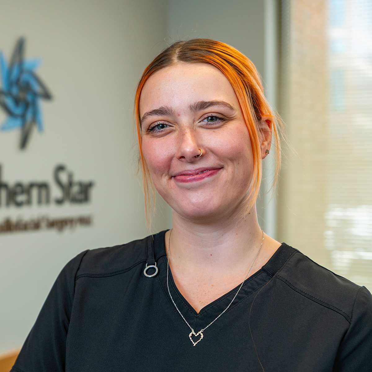 The image shows a person with red hair and a necklace smiling, wearing a black shirt with a white collar, standing against a background with a logo and text.
