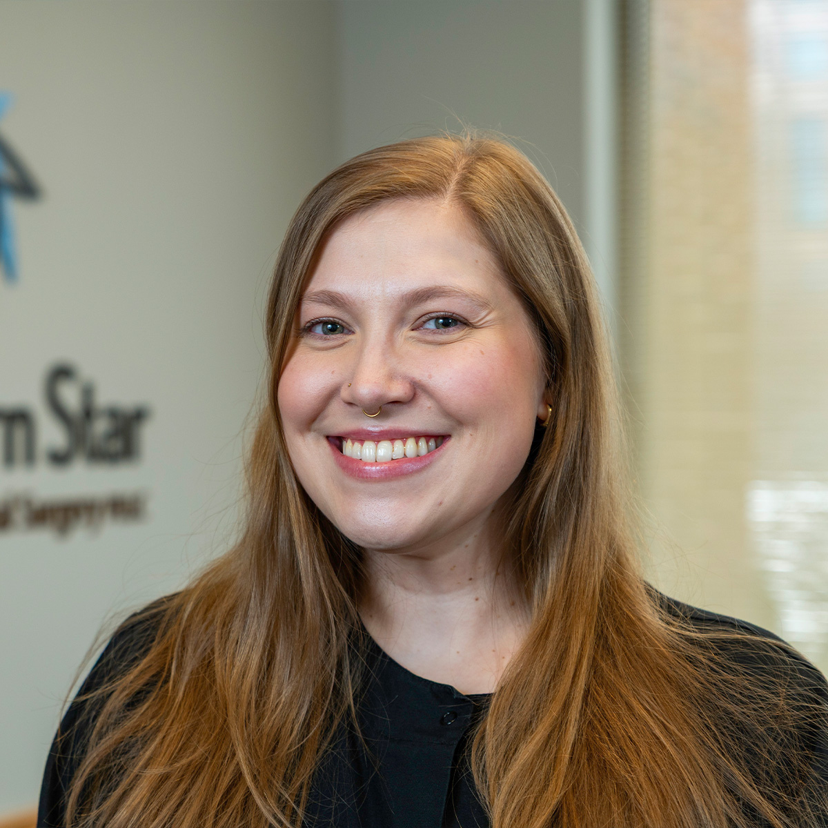 The image shows a smiling woman with long hair, wearing a dark top, standing in front of a wall with text and logos, including New Star .